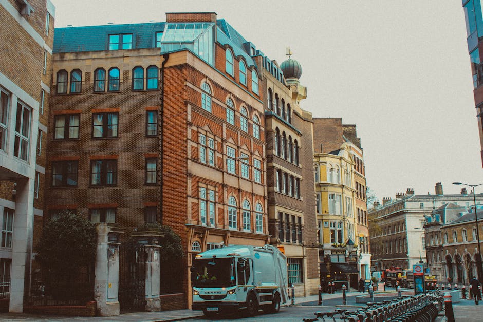 The image shows a street scene in West Hampstead, featuring multi-storey buildings with a mix of brick and ornate facades. A small delivery van with the branding of West Hampstead Man and Van is parked on the pavement, ready for home relocation or furniture transport. Nearby, there are several cardboard boxes and packing materials positioned near the building entrance, indicating ongoing packing or loading activities as part of a house removal. The scene includes a trolley or dolly, which may be used for carrying heavy items, and a vehicle loading area where items are being moved onto or from the van. The environment is lit with natural daylight, with historic architecture visible in the background and a clear street scene with bicycles secured along the sidewalk. This visual representation aligns with professional moving services and the logistical process involved in packing, loading, and transporting belongings during a house move in West Hampstead, adhering to Camden Council waste rules and regulations for waste disposal and skip usage.
