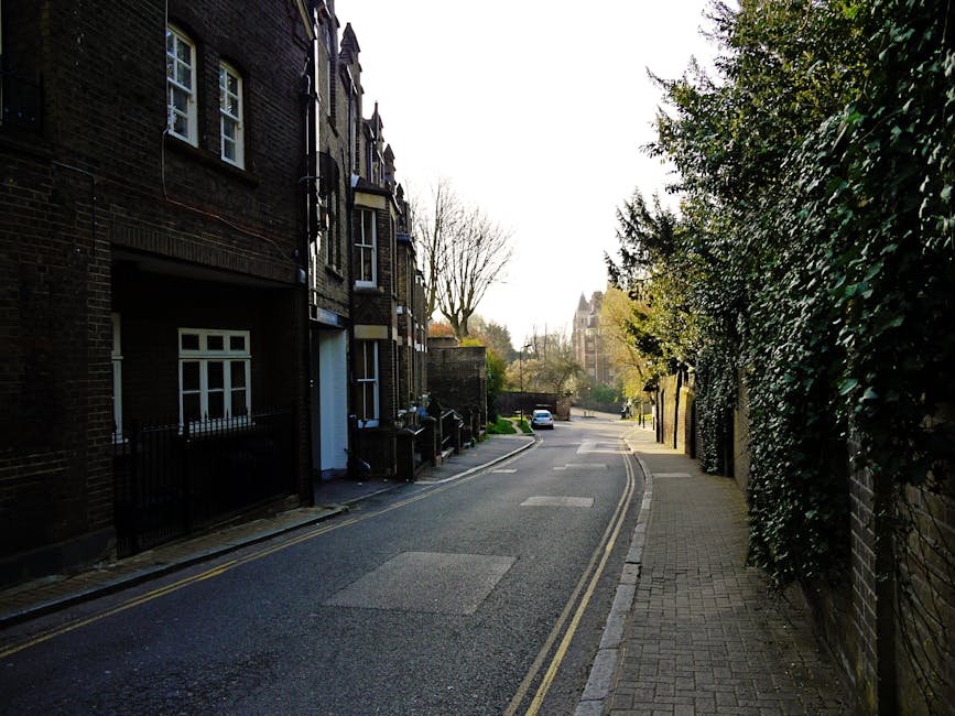 The image depicts a quiet residential street in West Hampstead during daytime, with a row of multi-storey brick houses on the left, featuring sash windows and front doors set back from the pavement. On the right, there is a sidewalk lined with tall, leafy green bushes and trees, casting shadows on the pavement. The street has double yellow lines along the edge, indicating parking restrictions, and is mostly clear with a few parked cars visible in the distance. The road appears to be in good condition, with some minor patches on the surface, and the scene is illuminated by natural daylight, creating a calm and orderly atmosphere suitable for home relocation and furniture transport activities. West Hampstead Man and Van, a professional removals company, may use this type of environment for loading or unloading household items, which are typically packed into cardboard boxes, wrapped furniture, and transported using vans, trolleys, and securing straps as part of a comprehensive packing and moving process. The surroundings reflect typical urban residential logistics relevant to Camden Council waste and skip rules for West Hampstead removals.