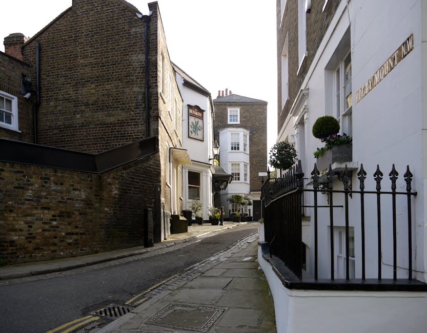 A narrow residential street in West Hampstead with a gentle incline paved with grey stone slabs, flanked by historic buildings made of brick and stucco. On the right, a white building features a small