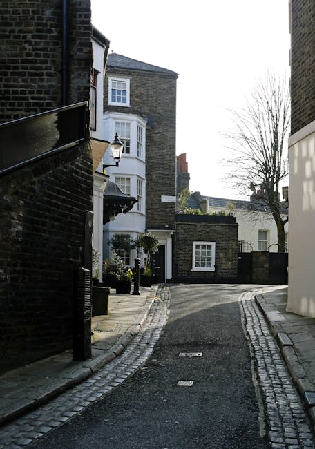A narrow residential street with cobbled paving and a slight incline, flanked by brick and stucco houses with bay windows and front gardens. On the left, a black metal railing runs alongside a building, with a street lamp mounted on the wall. At the end of the street, a tree with bare branches is visible against the sky, indicating a period of late autumn or winter. The scene is well-lit with natural daylight, and the street appears to be used temporarily for loading or unloading during a house removal. The setting reflects typical urban surroundings for home relocation services near Iverson Road, with potential access constraints for large furniture or moving vans. West Hampstead Man and Van would manage the logistics of furniture transport along this constrained route, considering the limited space and narrow access during the loading process.