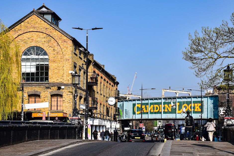 A street scene in West Hampstead featuring a large brick building with arched windows and a clock mounted on a pole. In the foreground, there are pedestrians walking along the pavement, some carrying shopping bags, and some waiting near a black fence. A vehicle, possibly a van used for removals, is partially visible on the left side. Overhead, a railway bridge painted with a green and yellow 'Caddon Lock' sign spans the street, with train tracks and electrical lines above. The environment is well-lit with clear blue skies, and a few trees with sparse foliage are visible on the right side of the image. The scene captures a typical urban setting suitable for home relocation or furniture transport activities, reflecting an active moving environment that could involve packing and loading procedures associated with house removals, as handled by companies like West Hampstead Man and Van.