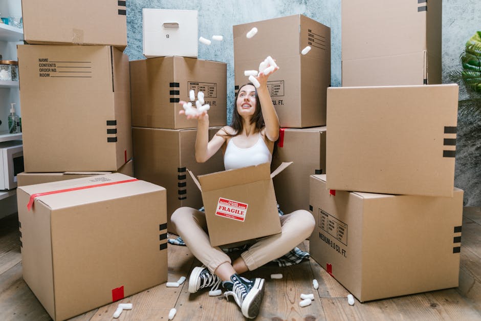 A young woman sits cross-legged on a wooden floor inside a property, surrounded by a large collection of packed cardboard boxes of various sizes, some sealed with red tape, and with labels indicating contents and room information. She is wearing casual clothing including a white tank top, beige pants, and sneakers, and is smiling while tossing packing foam peanuts into the air. One of the boxes she is sitting in has a red 'Fragile' sticker, suggesting careful handling for moving purposes. The background includes a blue textured wall and a partially visible green plant in the corner. Outside the property, a moving van with an open rear door is visible, indicating the ongoing process of home relocation, with furniture and moving equipment possibly nearby. The scene captures a moment during packing and furniture transport preparation, typical of professional removals services offered by West Hampstead Man and Van, supporting efficient packing and moving logistics.
