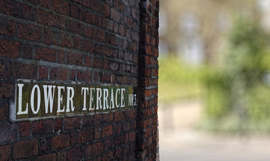 A close-up view of a brick wall displaying a street sign that reads 'LOWER TERRACE NW3' with white letters on a green background, mounted on the corner of a residential building in West Hampstead. In the background, a moving process is visible with a partially loaded or unloaded van parked outside, showing several cardboard boxes wrapped in plastic and brown packing paper. The boxes are placed on the pavement near the entrance, with some being carried or prepared for loading, indicating a home relocation or furniture transport scenario. The natural lighting highlights the textured brickwork and the organized arrangement of packing materials, consistent with professional removals by West Hampstead Man and Van, supporting services such as packing and furniture transport related to the West Hampstead removals on West End Lane NW6.
